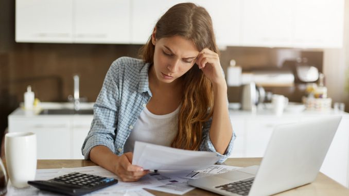 Attractive young housewife wearing shirt at home studying gas and electricity bills, checking calculations, looking at sheet of paper in her hands with serious and focused expression on her face zadłużenie